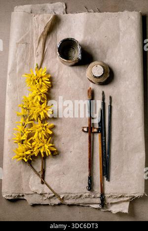 Flat lay composition with yellow forsythia branch vintage paintbrushes and ink container on handmade mulberry paper. Art supplies for painting or call Stock Photo