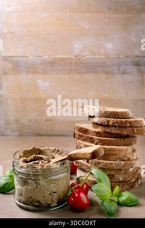 Homemade liver pate in glass jar with wooden spoon stack sliced whole grain bread fresh cherry tomatoes and basil leaves on rustic background Stock Photo
