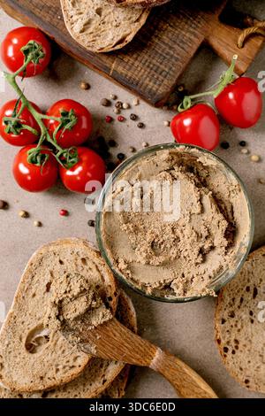 Homemade liver pate bowl with fresh bread slices ripe cherry tomatoes on vine and scattered peppercorns. Rustic wooden cutting board background. Tradi Stock Photo