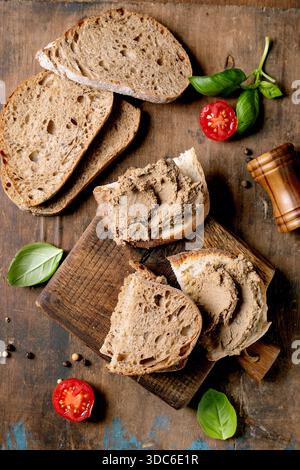 Sliced sourdough bread with liver pate spread on wooden cutting board. Fresh basil leaves and cherry tomatoes. Rustic wooden background. Homemade brea Stock Photo
