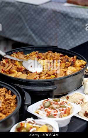 Appetizing dish of fried potatoes with corn bell peppers mushrooms in black cooking pan. Metal spoon in pan. Street food with side dishes visible in f Stock Photo
