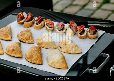 Delicious assortment appetizers: beef crostini topped with roasted peppers and golden baked empanadas on baking paper. Perfect catering food for party Stock Photo