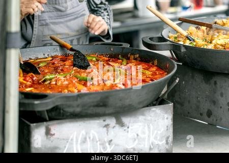 Chef in apron preparing red vegetable stew in large cast iron skillet. Tomato sauce with bell peppers cooking on stove while second pan contains stirf Stock Photo