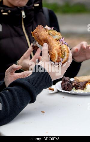 Person holding crispy sandwich with meat filling in paper wrapper. Street food outdoors. Blurred background with another plate of food on table. Stock Photo