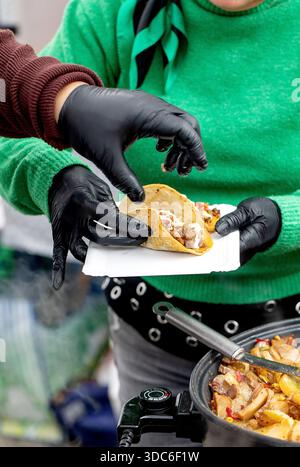 Person in green sweater and black gloves serving fresh taco from street food cart. Closeup of hands holding Mexican food with meat filling and sauce o Stock Photo