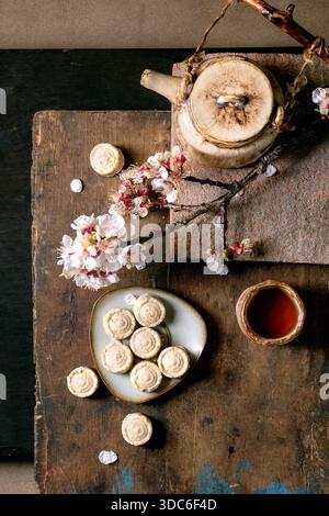 Traditional ceramic teapot with sesame cream candies on rustic wooden table decorated with flowering cherry blossom branches. Cup of tea completes atm Stock Photo