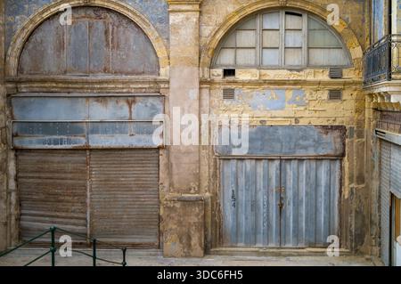 Weathered facades of old shops stand side-by-side. The left shop has a rusty roll-up door, while the right has a metal door with graffiti. Both are cl Stock Photo