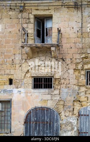 Weathered facade of an old building, possibly in Malta. The architecture shows a balcony with broken doors, barred windows, and arched doorways, hinti Stock Photo