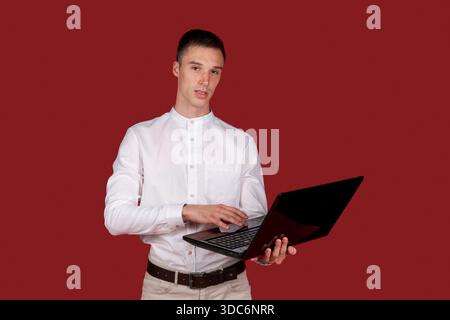 Portrait attractive focused cheerful brunette guy using laptop e-commerce project in white shirt isolated on bright red color background || Model released Stock Photo