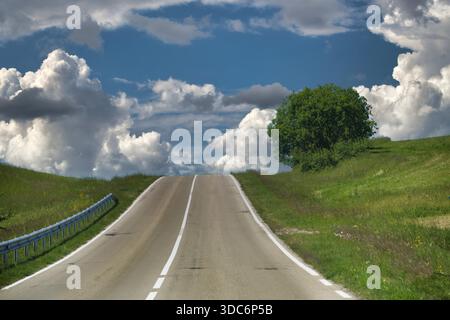 A quiet country road climbs a grassy hill, flanked by a guard rail and a lone tree on the right. Puffy clouds fill a blue sky, evoking freedom and open landscapes. Stock Photo