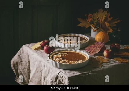 Traditional homemade autumn pumpkin pies for Thanksgiving or Halloween dinner served in ceramic dish with yellow autumn leaves, pumpkins and apples on linen table cloth. Dark rustic style Stock Photo