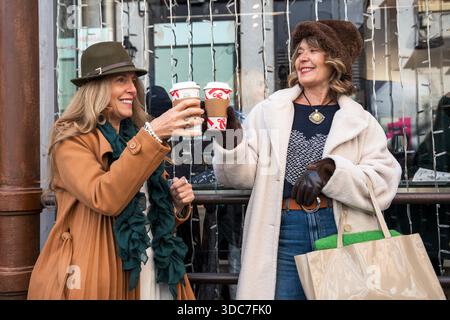 Two friends enjoy a cheerful moment, holding cups of warm beverages while standing outside a festive shop. Their smiles radiate joy as they celebrate Stock Photo