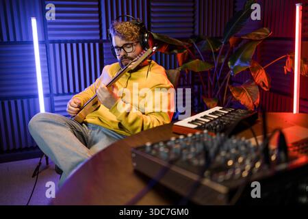 A focused male musician fine-tunes a guitar against the backdrop of a digital sound editing setup in a contemporary recording studio environment. Stock Photo