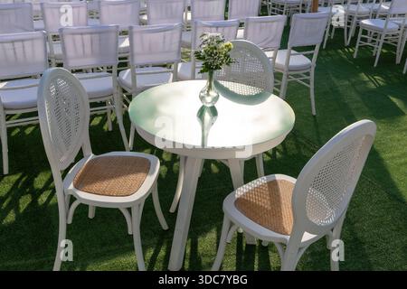 Round cafe table with small flower vase and three white chairs on green turf beside rows of conference seating in bright afternoon light suited for event breaks or networking space  Stock Photo