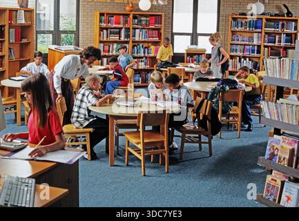 Students are seated at tables in a library. Some read, others write in notebooks. A teacher helps a few students. Books are on shelves around them. Stock Photo