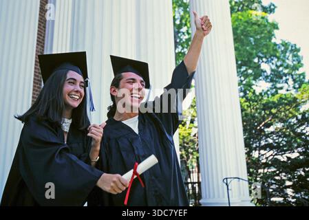 Happy and smiling male and female college graduates. One holds a diploma while the other gives a thumbs up. They are dressed in graduation gowns. Stock Photo
