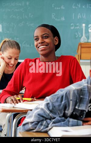 Smiling African-American female high school student paying attention in class. One student is smiling while writing, and another student in the backgr Stock Photo