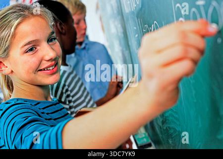 Female call Kasian student in a classroom activity where she writes on a chalkboard. They use chalk to express ideas while teachers guide them. Stock Photo