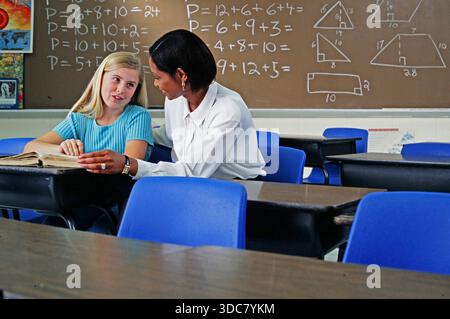 A student and a teacher engage in a math lesson, discussing problems and solutions at a desk in a classroom. Focus is on learning and teaching in the Stock Photo
