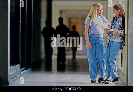 Two girls stand in a school hallway talking and smiling with books in their hands. Other students walk in the background. The setting is bright and li Stock Photo