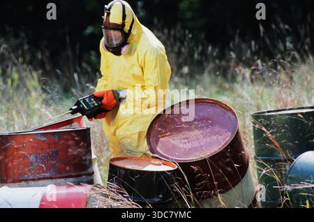 A worker in a yellow protective suit gathers data from barrels in a  toxic waste dump. The task takes place during the day with tall grass surrounding Stock Photo