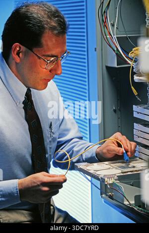 A technician is focused on making connections inside a server rack. He is wearing glasses and a tie. Wires are visible as he handles computer componen Stock Photo