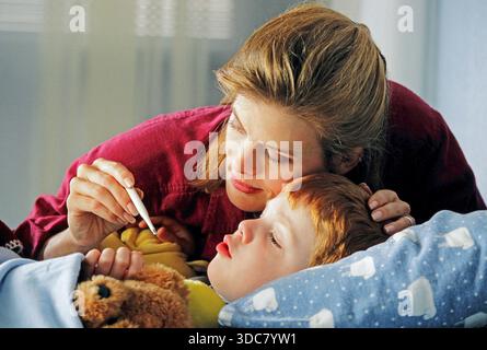 A mother comforts her sick child in bed. She uses a thermometer to check his temperature. The child looks tired and is lying under a blanket with a te Stock Photo