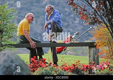 Man and eldwely father  sit on a bench at a golf course. They are laughing and talking while surrounded by colorful plants and a golf bag nearby. It i Stock Photo