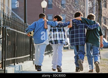 Four  teen boys  walk side by side on a sidewalk in a city. They wear casual clothes and one of them holds a soccer ball. They seem to enjoy each othe Stock Photo