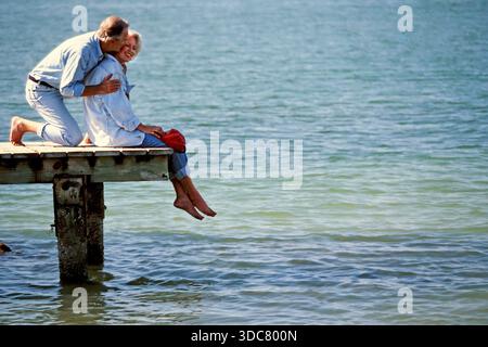 Senior couple sit on a wooden dock above the water. They smile and share a moment while their feet dangle above the surface. It is sunny and they look went Stock Photo