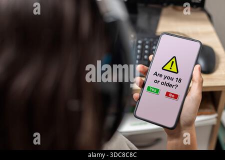 An unrecognizable female teen is holding a mobile phone with legal age verification displaying on its screen Stock Photo