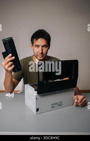 Vertical portrait of computer technician replacing toner cartridge in laser printer during maintenance service, holding new cartridge and looking at Stock Photo