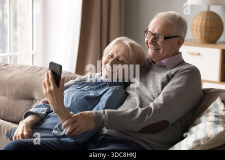 Smiling senior family couple cuddle on couch using smartphone Stock Photo
