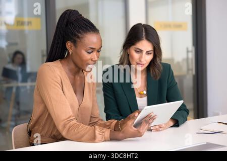Two engaged multiethnic female colleagues using tablet at workplace Stock Photo