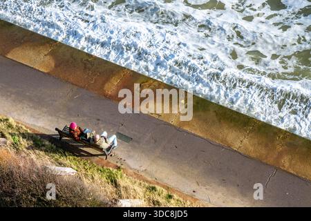 Elderly couple sitting on a bench at the seaside with waves breaking onto the sea wall, couple watching the waves at the seaside at Shanklin, IOW, UK Stock Photo
