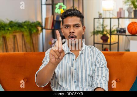Indian man at home waves finger no no to camera and shows refusal and disagreement with stern mood. Arabian guy on sofa shakes head denies request points finger and shows unhappy sad reject reaction Stock Photo