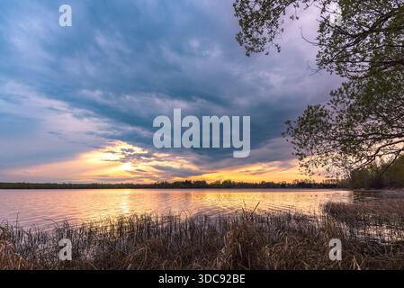 Scenic summer evening view of the sun setting behind dark storm clouds, reflecting in Lacombe Lake at the Lacombe Lake boat launch in Alberta, Canada. Stock Photo