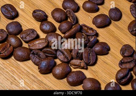 Numerous dark brown roasted coffee beans are spread randomly over a warm, light-colored wooden table, emphasizing their texture. Stock Photo