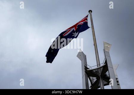 The Australian national flag flutters in the wind from a tall flagpole, rising above a modern metallic pylon under a cloudy sky. Stock Photo