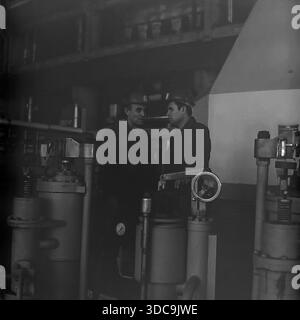 Slaviansk, Ukrainian SSR, USSR - 1981: Archival industrial photograph featuring Deputy Rypalenko discussing production issues with a colleague on the factory floor. Both men wear safety helmets and work clothes while standing near complex heavy machinery with valves and pressure gauges. In the background, a control panel is visible. This image captures the working atmosphere and technical collaboration in the industrial sector of the peaceful Donbass region during the late Soviet era. Stock Photo