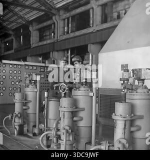 Slaviansk, Ukrainian SSR, USSR - 1981: Archival industrial photograph featuring Deputy Rypalenko discussing production issues with a colleague on the factory floor. Both men wear safety helmets and work clothes while standing near complex heavy machinery with valves and pressure gauges. In the background, a control panel is visible. This image captures the working atmosphere and technical collaboration in the industrial sector of the peaceful Donbass region during the late Soviet era. Stock Photo