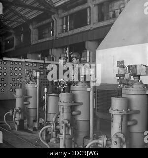 Slaviansk, Ukrainian SSR, USSR - 1981: Archival industrial photograph featuring Deputy Rypalenko discussing production issues with a colleague on the factory floor. Both men wear safety helmets and work clothes while standing near complex heavy machinery with valves and pressure gauges. In the background, a control panel is visible. This image captures the working atmosphere and technical collaboration in the industrial sector of the peaceful Donbass region during the late Soviet era. Stock Photo