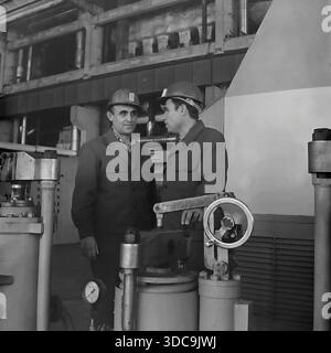 Slaviansk, Ukrainian SSR, USSR - 1981: Archival industrial photograph featuring Deputy Rypalenko discussing production issues with a colleague on the factory floor. Both men wear safety helmets and work clothes while standing near complex heavy machinery with valves and pressure gauges. In the background, a control panel is visible. This image captures the working atmosphere and technical collaboration in the industrial sector of the peaceful Donbass region during the late Soviet era. Stock Photo