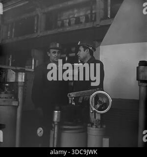 Slaviansk, Ukrainian SSR, USSR - 1981: Archival industrial photograph featuring Deputy Rypalenko discussing production issues with a colleague on the factory floor. Both men wear safety helmets and work clothes while standing near complex heavy machinery with valves and pressure gauges. In the background, a control panel is visible. This image captures the working atmosphere and technical collaboration in the industrial sector of the peaceful Donbass region during the late Soviet era. Stock Photo