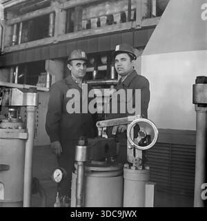 Slaviansk, Ukrainian SSR, USSR - 1981: Archival industrial photograph featuring Deputy Rypalenko discussing production issues with a colleague on the factory floor. Both men wear safety helmets and work clothes while standing near complex heavy machinery with valves and pressure gauges. In the background, a control panel is visible. This image captures the working atmosphere and technical collaboration in the industrial sector of the peaceful Donbass region during the late Soviet era. Stock Photo