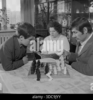 Slaviansk, Ukrainian SSR, USSR - 1981: Archival lifestyle photograph capturing a Soviet family evening. A father with a mustache plays chess with his son at a table, while the mother reads a magazine in the background. The room features a typical late-Soviet interior, including a polished wall unit ('stenka') filled with crystal and books. This scene reflects the popularity of chess and domestic stability in the peaceful Donbass region during the late Soviet era. Stock Photo