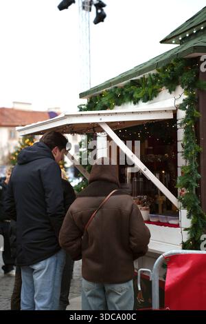 people in winter attire visit a festive market stall decorated with greenery and lights, set against a urban backdrop with cobblestone path and christ Stock Photo