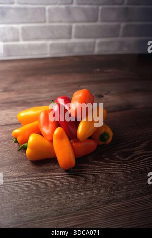 a vibrant assortment of mini bell peppers in vivid red, orange, and yellow hues arranged on a rustic wooden table against a textured stone brick wall Stock Photo