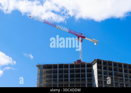 High-rise residential building under construction with a large red and white tower crane on top against a bright blue sky with clouds Stock Photo