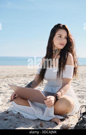 Woman in white summer dress sits on the beach with laptop with eyes closed, relaxing, enjoy summer sun, distracted from freelance work or studying Stock Photo